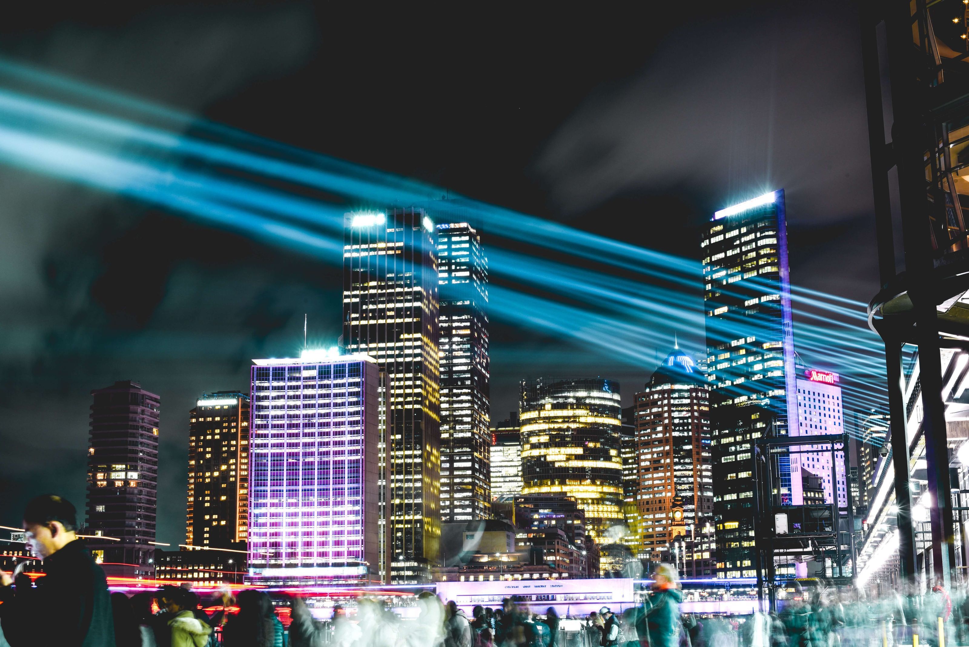 This photo shows a lively cityscape at night. Buildings are colorfully lit up against the night sky and bright beams cross the photo. 